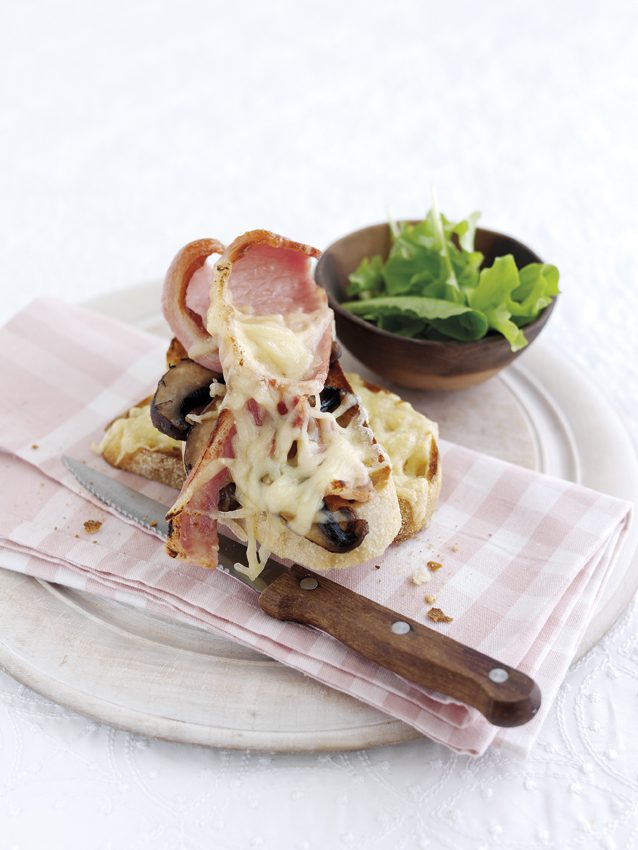 Two bacon rashers covered in melted gruyere cheese with mushroom. Served on a plate with knife on side.