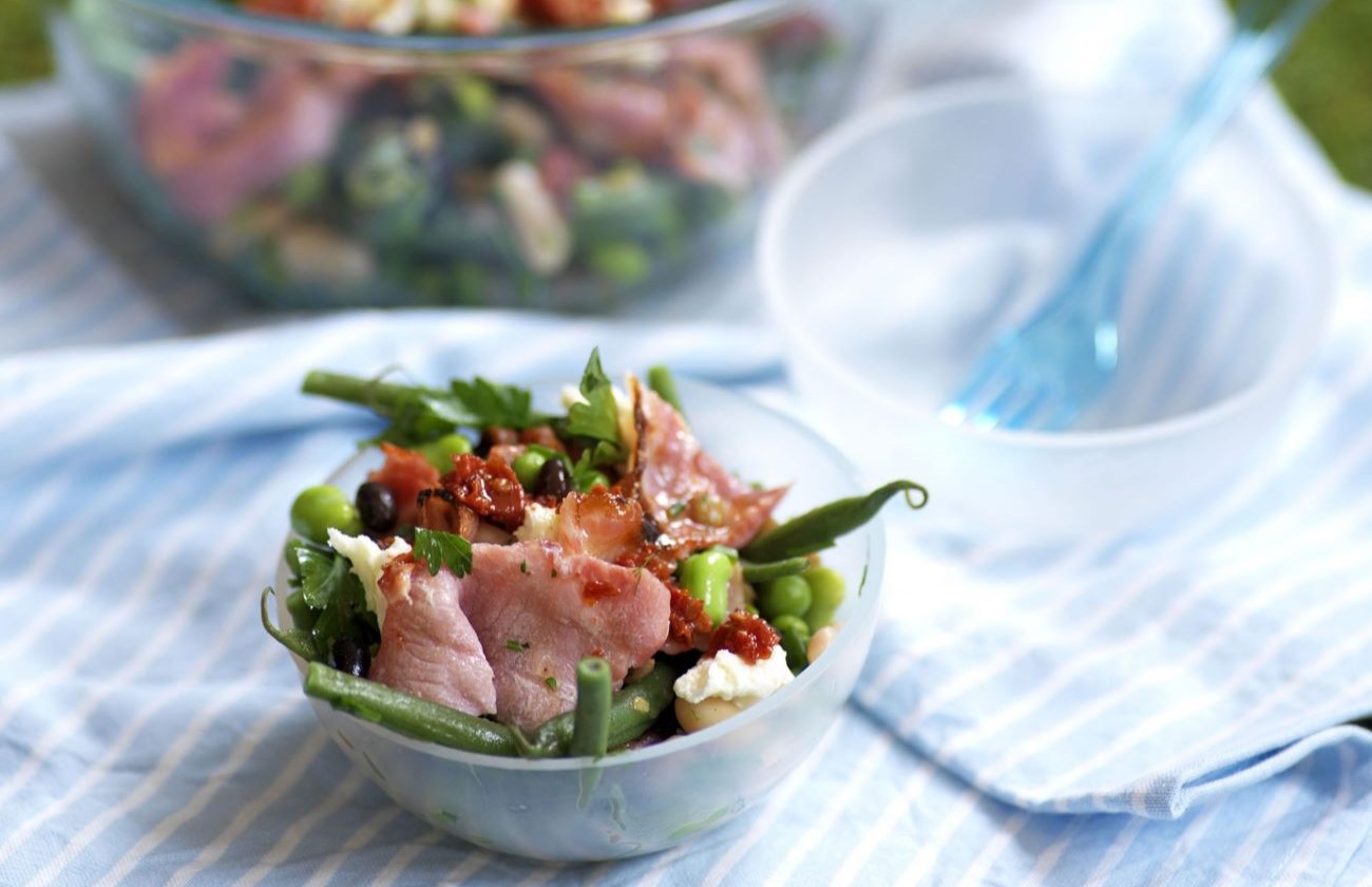 Small white bowl on white table cloth with bacon bean and herb salad, main mixing bowl in background