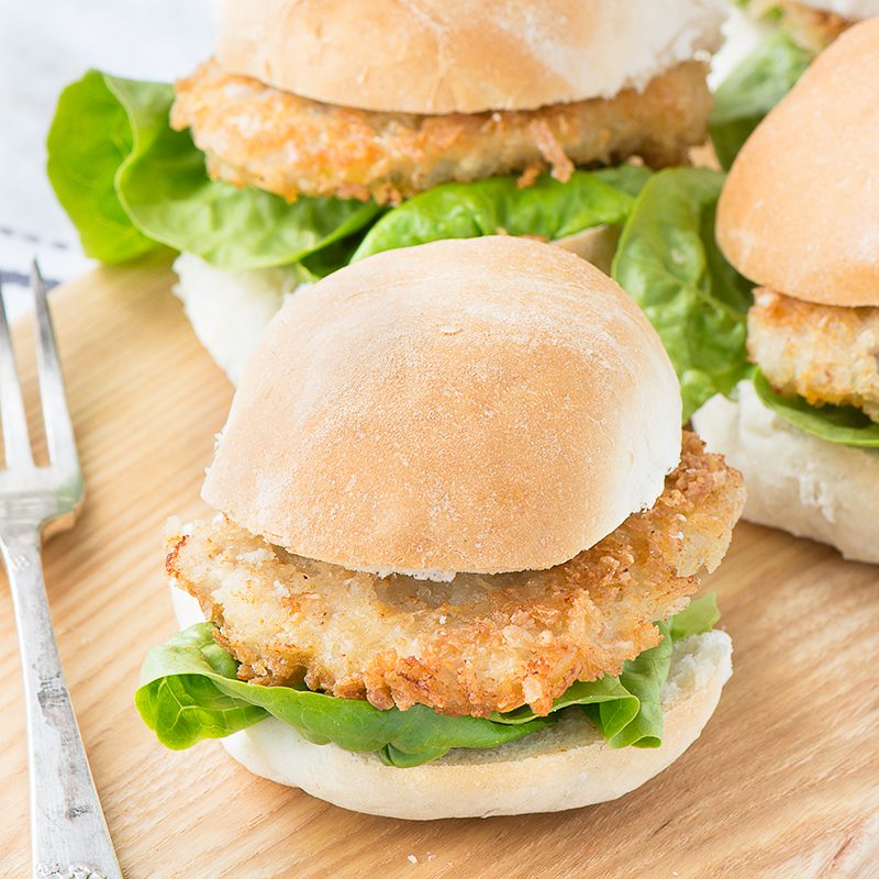 Crispy coated pork medallion in between two burger buns with lettuce. On a wooden chopping board.
