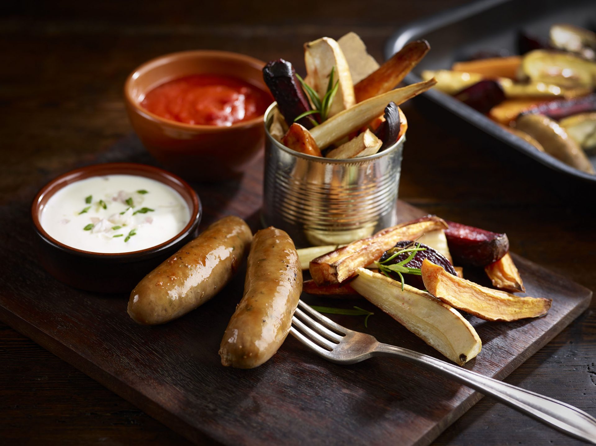 Two pork sausages served on a black platter with dips and roasted vegetables